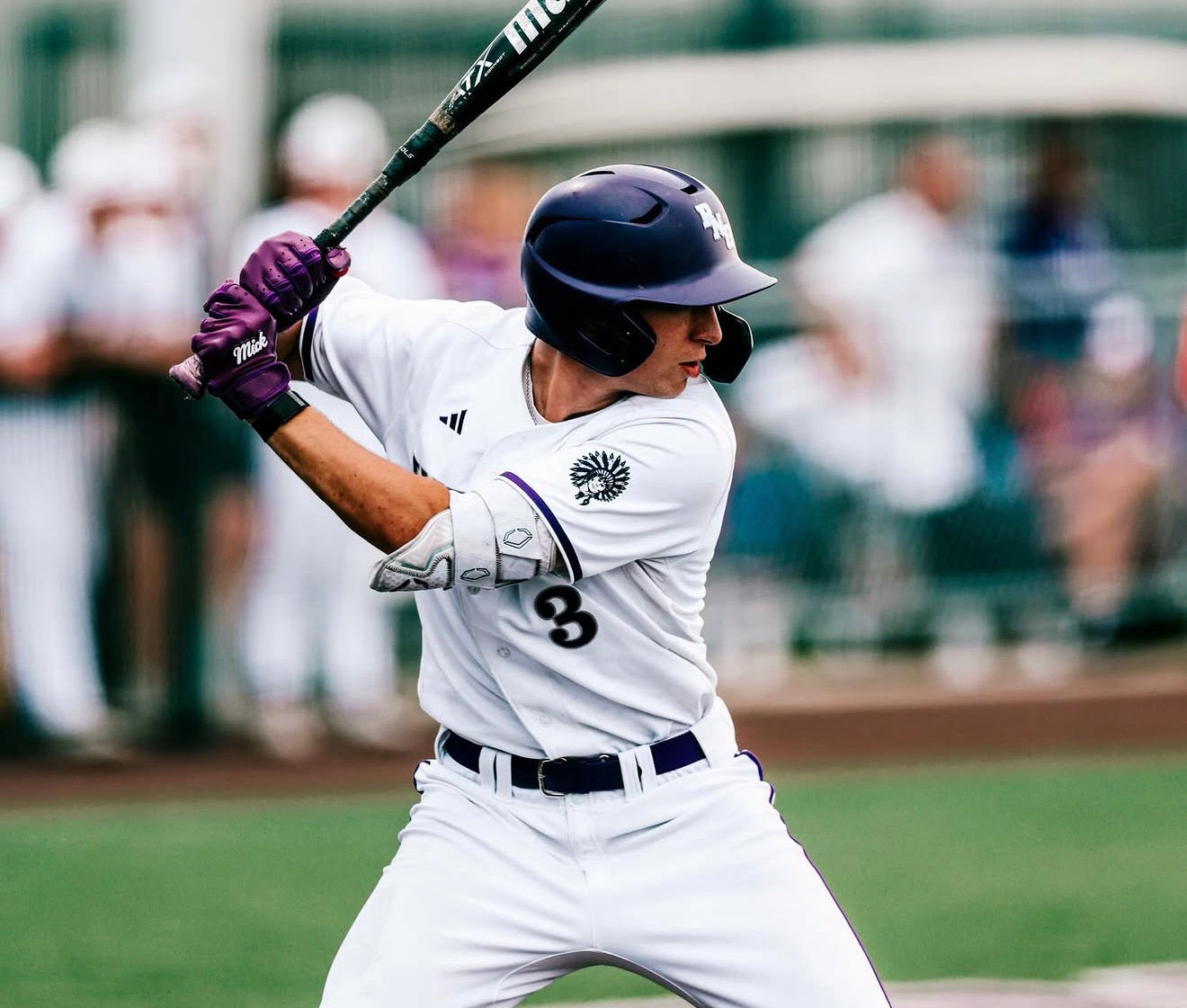 Baseball player in a white uniform with purple helmet and bat, ready to swing on a baseball field.